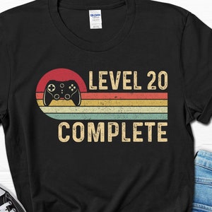 May include: Black t-shirt with a retro-style graphic of a video game controller and the text "LEVEL 20 COMPLETE".