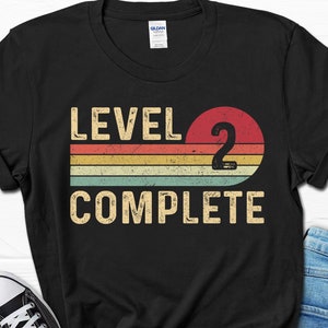 May include: Black t-shirt with a vintage-style graphic that says "Level 2 Complete" with a large red number 2 and colorful stripes.