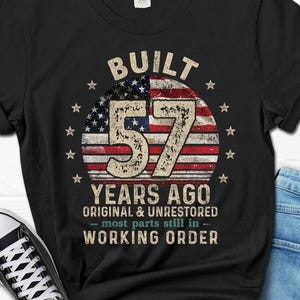 May include: Black t-shirt with a graphic celebrating a 57th birthday. The design features the text "BUILT 57 YEARS AGO ORIGINAL & UNRESTORED - most parts still in - WORKING ORDER" with an American flag background.