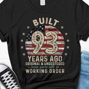 May include: Black t-shirt with a distressed design featuring the text "BUILT 93 YEARS AGO" with an American flag graphic. The design includes stars and the phrase "ORIGINAL & UNRESTORED - MOST PARTS STILL IN - WORKING ORDER."