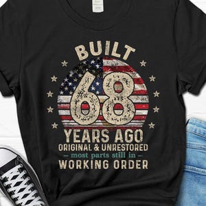 May include: Black t-shirt with a distressed design featuring the text "BUILT 68 YEARS AGO" above a circular American flag graphic. The text "ORIGINAL & UNRESTORED - MOST PARTS STILL IN - WORKING ORDER" is below the graphic.