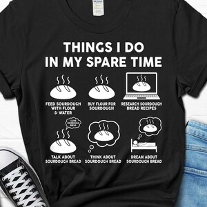 May include: A black t-shirt with white text that reads "Things I do in my spare time". The shirt features a series of illustrations depicting activities related to sourdough bread, including feeding sourdough, buying flour, researching recipes, talking about sourdough, thinking about sourdough, and dreaming about sourdough.