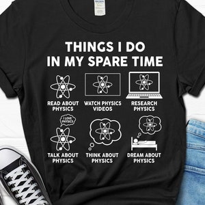 May include: Black t-shirt with white text that reads "Things I do in my spare time". The text is surrounded by illustrations of atoms and phrases related to physics, such as "Read about physics", "Watch physics videos", "Research physics", "Talk about physics", "Think about physics", and "Dream about physics".