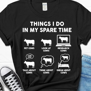 以下が含まれることがあります： 「Things I do in my spare time」という白い文字が書かれた黒いTシャツ。テキストは、牛を描いた6つの白いアイコンで囲まれています。アイコンは、なでられている牛、見られている牛、ラップトップで調査されている牛、話されている牛、考えられている牛、夢に見られている牛を表しています。