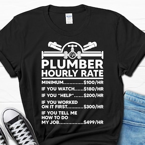 May include: Black t-shirt with a white graphic of a plumber's tools and the text "Plumber Hourly Rate". The graphic includes a pipe, wrench, and a valve. The text below the graphic lists different hourly rates for a plumber based on the level of assistance provided.  The rates are: $100/hr, $180/hr, $200/hr, $300/hr, and $499/hr.