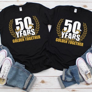 May include: Two black t-shirts with gold lettering and a gold laurel wreath design. The text on the shirts reads "50 Years We are Golden Together".