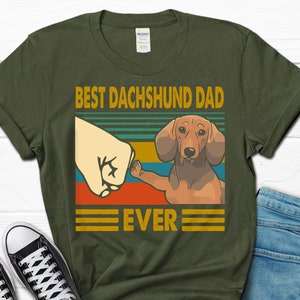 May include: Green t-shirt with a vintage-style graphic of a dachshund giving a fist bump. The text reads "Best Dachshund Dad Ever".