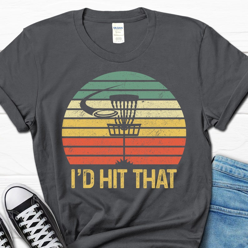 May include: A dark gray t-shirt with a vintage-style graphic of a disc golf basket and the text "I'd Hit That".