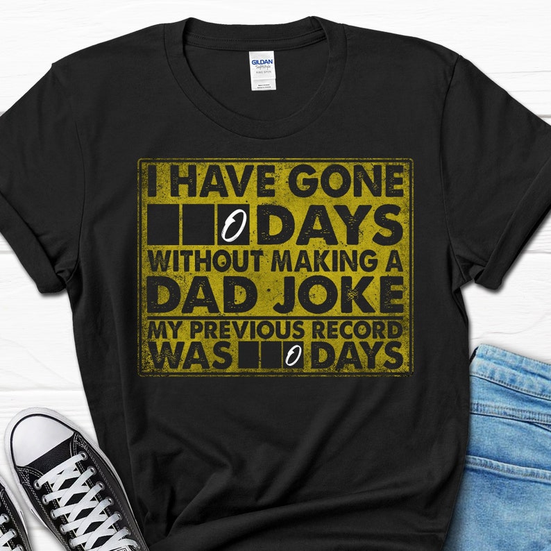 Op de afbeelding: Zwart T-shirt met een gele afbeelding waarop staat: "Ik ben 0 dagen zonder een vaders grapje gegaan. Mijn vorige record was 0 dagen."