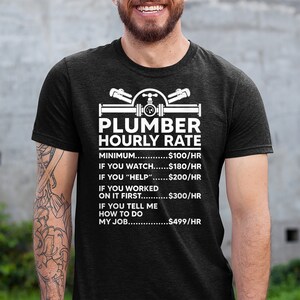 May include: A black t-shirt with a white graphic of a plumber's tools and the text "Plumber Hourly Rate". The graphic includes a pipe, wrench, and valve. The text below the graphic lists different hourly rates for a plumber based on the level of assistance provided. The rates are: $100/hr for minimum, $180/hr for watching, $200/hr for helping, $300/hr for working on it first, and $499/hr for telling the plumber how to do the job.