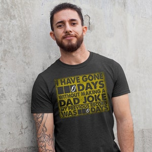 Op de afbeelding: Een donkergrijs T-shirt met een gele afbeelding waarop staat: "Ik ben 0 dagen zonder een vaders grap gegaan. Mijn vorige record was 0 dagen."
