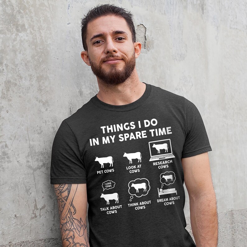 以下が含まれることがあります： 暗いグレーのTシャツに、白い文字で「Things I do in my spare time」と書いてあり、牛に関連する活動のリストが示されています。活動には、牛をなでる、牛を見る、牛について調べる、牛について話す、牛について考える、牛について夢を見るなどがあります。