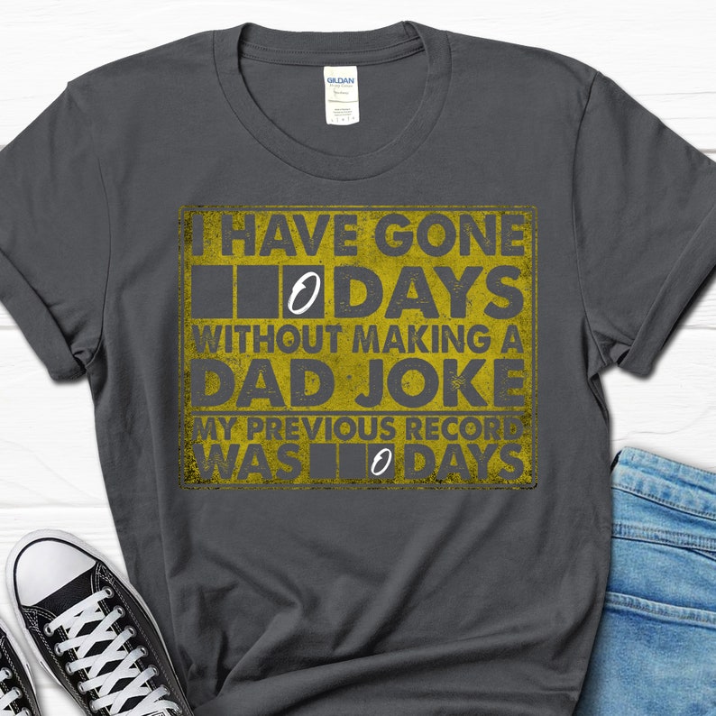 Op de afbeelding: Een donkergrijs T-shirt met een gele afbeelding waarop staat: "Ik ben 0 dagen zonder een vadersgrap gegaan. Mijn vorige record was 0 dagen."