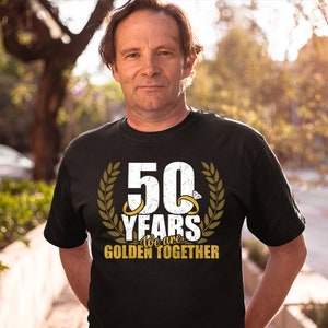 May include: A black t-shirt with a white graphic that says "50 Years We are Golden Together". The graphic features a large number 50 with two gold rings and a diamond, surrounded by a laurel wreath.