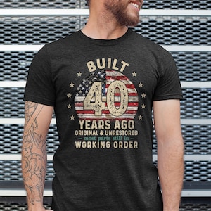 May include: A dark heather grey t-shirt with a distressed American flag design and the text "BUILT 40 YEARS AGO ORIGINAL & UNRESTORED - most parts still in - WORKING ORDER".