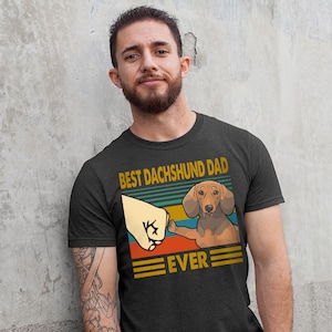 May include: A dark grey t-shirt with a vintage-style graphic of a dachshund giving a high five. The graphic says "Best Dachshund Dad Ever".