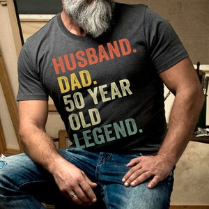 May include: A dark gray t-shirt with a vintage-style graphic that reads "Husband. Dad. 50 Year Old Legend."