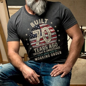 May include: A dark gray t-shirt with a vintage American flag design and the text "Built 70 Years Ago Original & Unrestored - most parts still in Working Order".