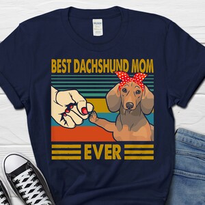 May include: Navy blue t-shirt with a retro-style graphic of a dachshund wearing a red bandana and the text "Best Dachshund Mom Ever".