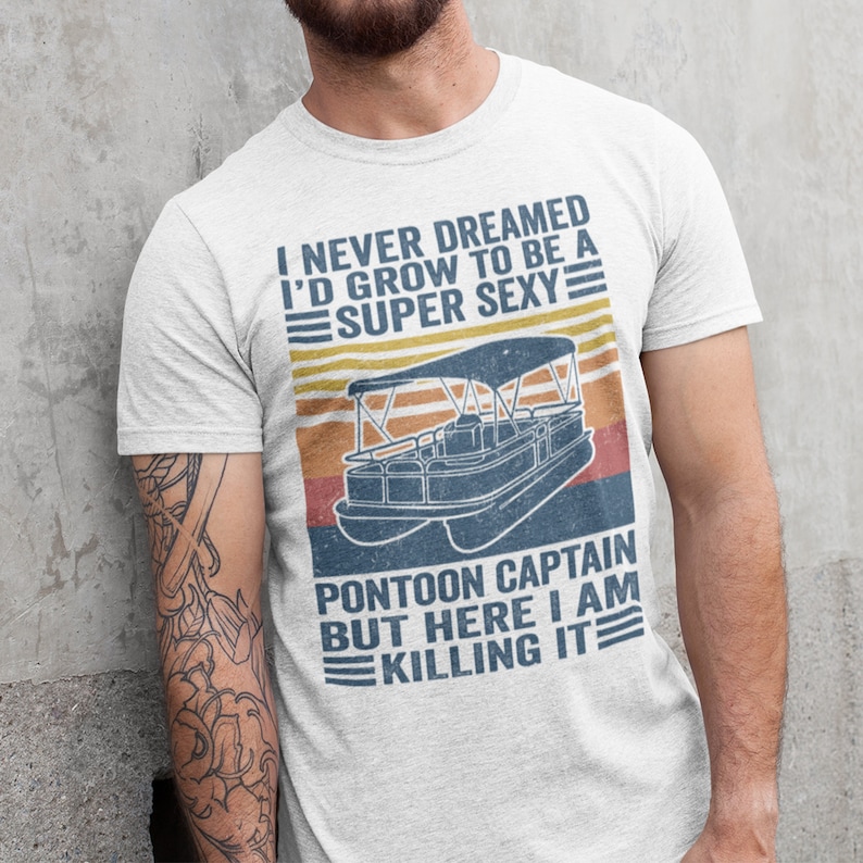 Pu&ograve; includere: Una maglietta bianca con una grafica in stile vintage di una barca pontone. Il testo sulla maglietta recita "I never dreamed I'd grow to be a super sexy pontoon captain but here I am killing it."