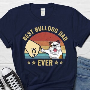 May include: Navy blue t-shirt with a vintage-style sunset graphic and the text "Best Bulldog Dad Ever" in white. A bulldog with a brown nose and ears is giving a thumbs up.