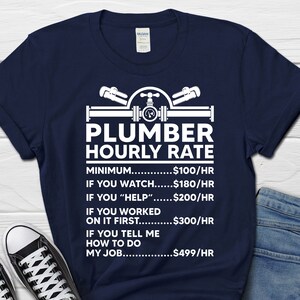 May include: A navy blue t-shirt with a white graphic of a plumber's tools and the text "Plumber Hourly Rate". The graphic includes a pipe, wrench, and a pressure gauge. The text below the graphic lists different hourly rates for a plumber based on the level of assistance provided, such as "Minimum $100/HR", "If you watch $180/HR", "If you "help" $200/HR", "If you worked on it first $300/HR", and "If you tell me how to do my job $499/HR".