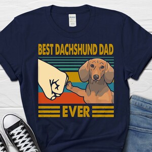 May include: Navy blue t-shirt with a retro-style graphic of a dachshund dog and the text "Best Dachshund Dad Ever".