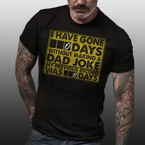 Op de afbeelding: Zwart T-shirt met een gele waarschuwingsbord-afbeelding met de tekst "Ik ben @ dagen zonder een vadersgrap gegaan. Mijn vorige record was @ dagen."
