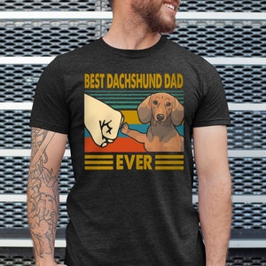 May include: A dark grey t-shirt with a vintage-style graphic of a dachshund giving a fist bump. The graphic says "Best Dachshund Dad Ever".