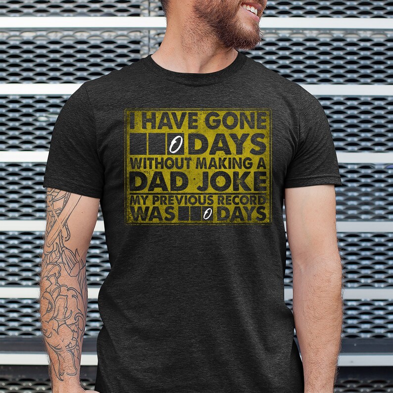 Op de afbeelding: Een donkergrijs T-shirt met een gele afbeelding waarop staat: "Ik ben 0 dagen zonder een vadersgrap gegaan. Mijn vorige record was 0 dagen."
