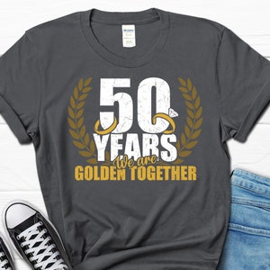 May include: A dark gray t-shirt with a gold graphic that says "50 Years We are Golden Together". The graphic features two gold rings with diamonds and a laurel wreath.