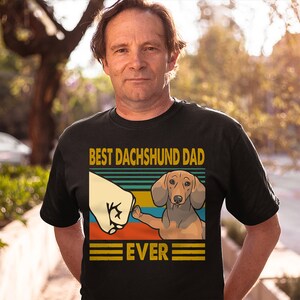 May include: A black t-shirt with a retro-style graphic of a dachshund giving a fist bump. The text "Best Dachshund Dad Ever" is printed on the shirt.