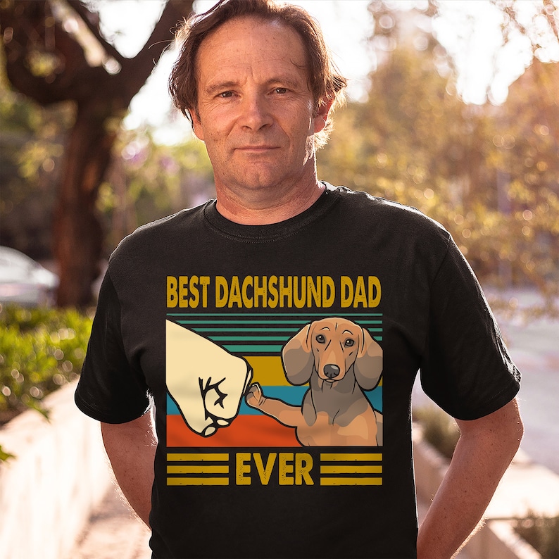 May include: A black t-shirt with a retro-style graphic of a dachshund giving a fist bump. The text "Best Dachshund Dad Ever" is printed on the shirt.