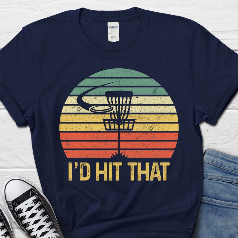 May include: A navy blue t-shirt with a vintage-style sunset graphic featuring a disc golf basket and the text "I'd Hit That".
