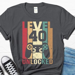 May include: A dark gray t-shirt with a vintage-style graphic that says "Level 40 Unlocked" with a retro video game controller in the center.