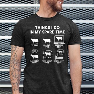 以下が含まれることがあります： 「Things I do in my spare time」というテキストと、牛に関連する活動のリストが書かれた濃いグレーのTシャツ。活動には、牛をなでる、牛を見る、牛を調べる、牛について話す、牛について考える、牛を夢見るなどがあります。