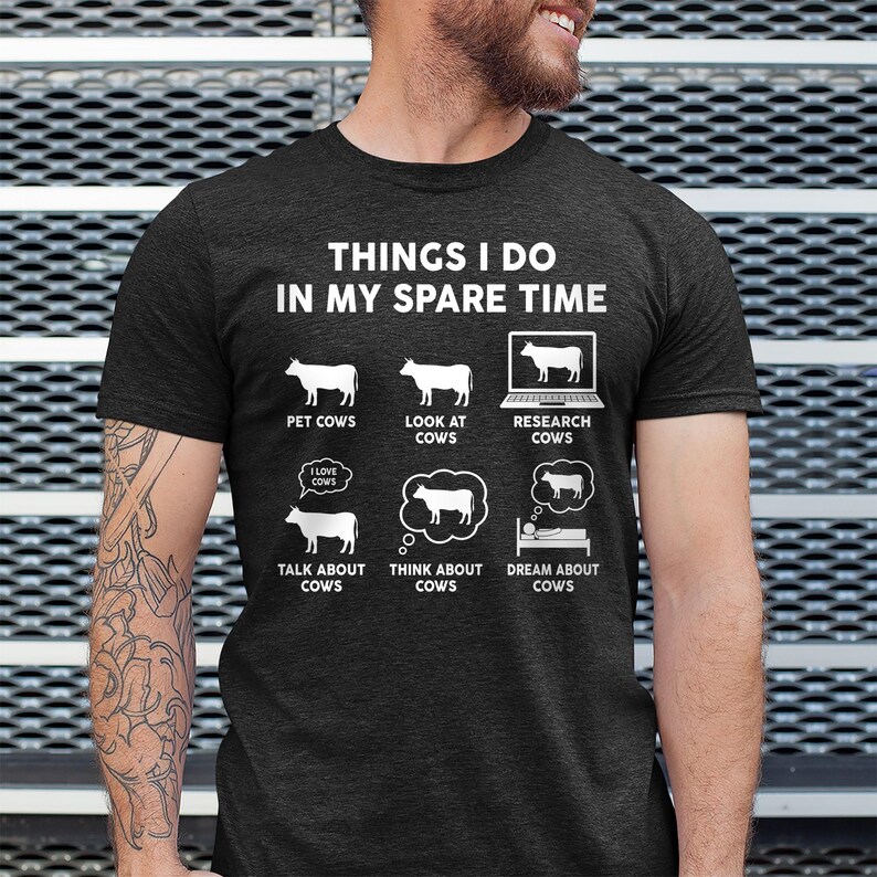 以下が含まれることがあります： 「Things I do in my spare time」というテキストと、牛に関連する活動のリストが書かれた濃いグレーのTシャツ。活動には、牛をなでる、牛を見る、牛を調べる、牛について話す、牛について考える、牛を夢見るなどがあります。