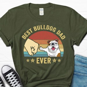 May include: Green t-shirt with a retro sunset graphic and the text "Best Bulldog Dad Ever". A bulldog paw is bumping fists with a hand.