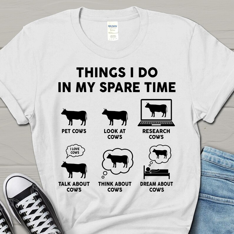 以下が含まれることがあります： 白いTシャツに黒い文字で「Things I do in my spare time」と書かれています。テキストの下には、牛のイラストが6つあり、それぞれにテキストが書かれています。イラストは、牛をなでている、牛を見ている、牛をラップトップで調べている、牛について話している、牛について考えている、牛を夢見ている様子を表しています。