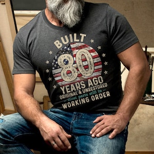 May include: A dark gray t-shirt with a vintage American flag design. The text "Built 80 Years Ago Original & Unrestored - most parts still in working order" is printed on the shirt.
