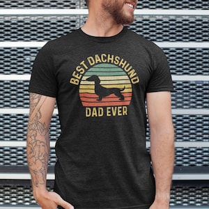 May include: A dark grey t-shirt with a vintage-style graphic of a dachshund in front of a sunset. The text "Best Dachshund Dad Ever" is printed below the graphic.