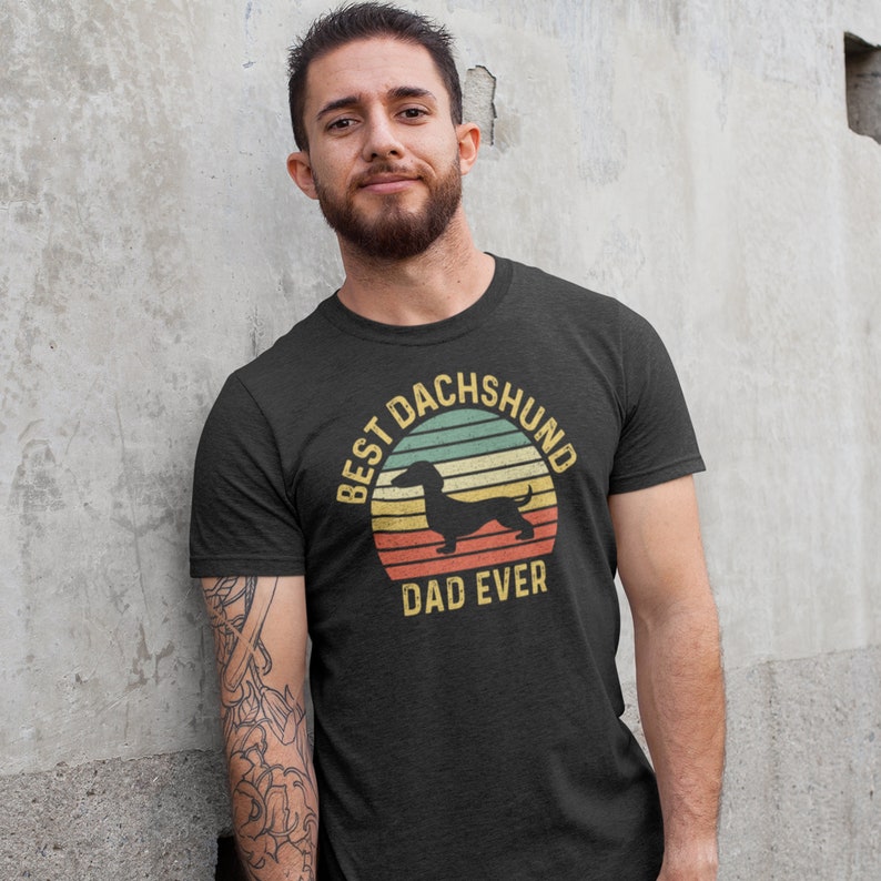 May include: A dark grey t-shirt with a vintage-style graphic of a dachshund silhouette in front of a sunset. The text "Best Dachshund Dad Ever" is printed below the graphic.