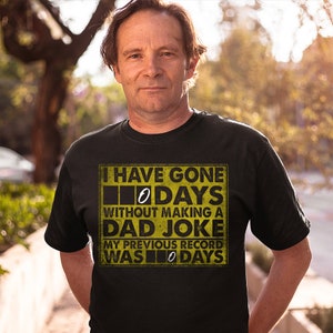 Op de afbeelding: Een zwart T-shirt met een gele afbeelding waarop staat: "Ik ben 0 dagen zonder een vaders grapje gegaan. Mijn vorige record was 0 dagen."