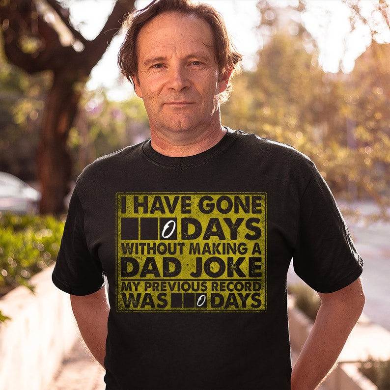 Op de afbeelding: Een zwart T-shirt met een gele afbeelding waarop staat: "Ik ben 0 dagen zonder een vaders grapje gegaan. Mijn vorige record was 0 dagen."