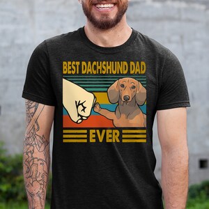 May include: A black t-shirt with a vintage-style graphic of a dachshund dog giving a fist bump. The text on the shirt reads "Best Dachshund Dad Ever".