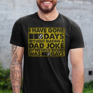 Op de afbeelding: Een zwart T-shirt met een gele afbeelding waarop staat: "Ik ben 0 dagen zonder een vaders grapje gegaan. Mijn vorige record was 0 dagen."
