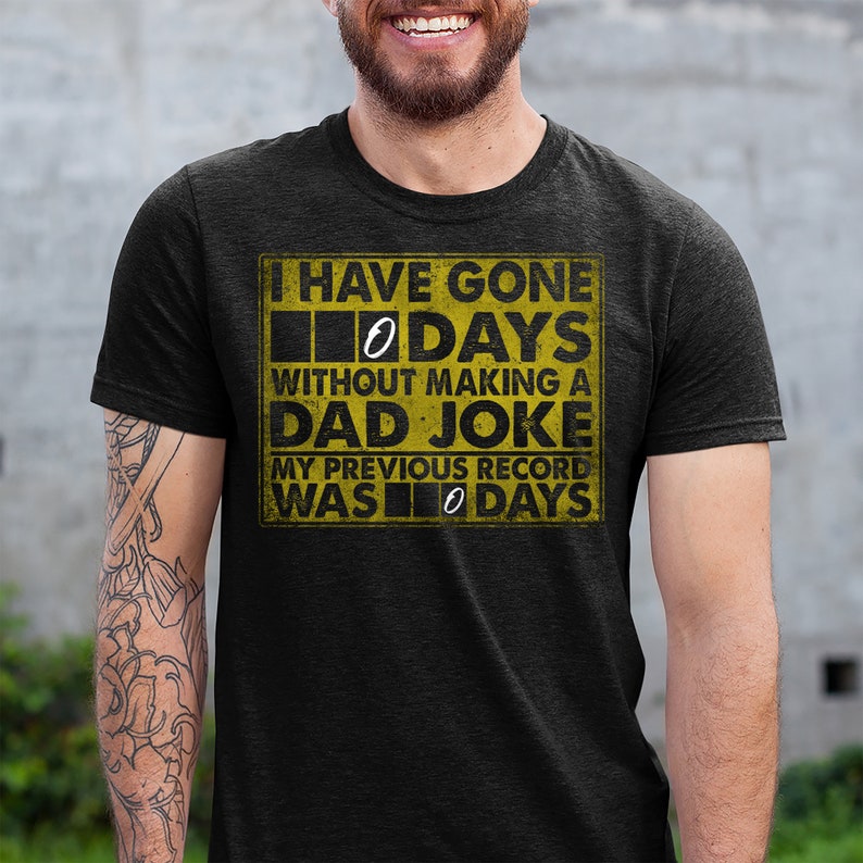 Op de afbeelding: Een zwart T-shirt met een gele afbeelding waarop staat: "Ik ben 0 dagen zonder een vaders grapje gegaan. Mijn vorige record was 0 dagen."