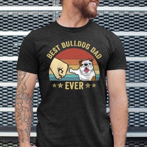 May include: A dark grey t-shirt with a vintage-style graphic that says "Best Bulldog Dad Ever". The graphic features a bulldog pawing at a fist in front of a sunset.