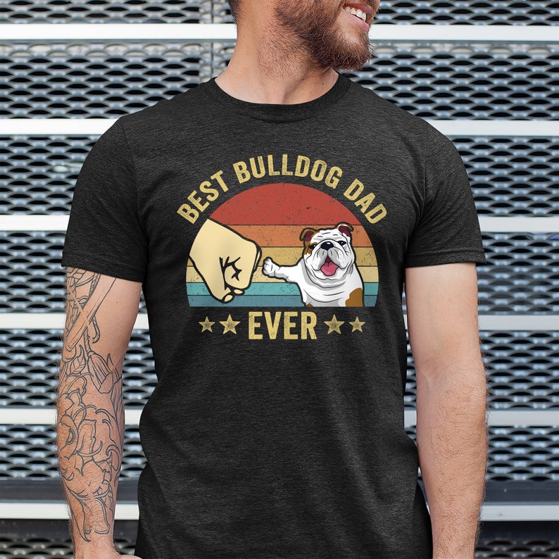 May include: A dark grey t-shirt with a vintage-style graphic that says "Best Bulldog Dad Ever". The graphic features a bulldog pawing at a fist in front of a sunset.