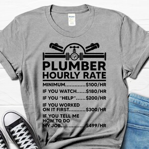 May include: Gray t-shirt with a black graphic of a plumber's tools and the text "Plumber Hourly Rate". The graphic includes a list of hourly rates for different levels of assistance, such as "Minimum", "If you watch", "If you "help"", "If you worked on it first", and "If you tell me how to do my job".